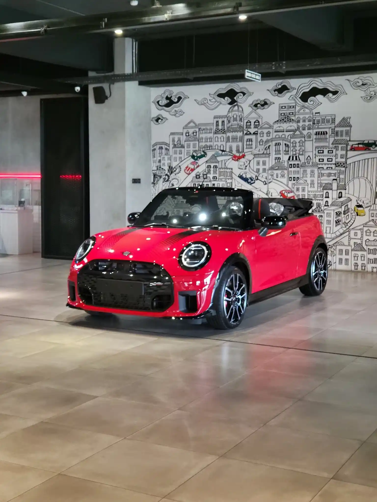 Mini Cooper bekas warna merah dipajang di showroom bergaya industrial dengan latar mural artistik hitam putih.